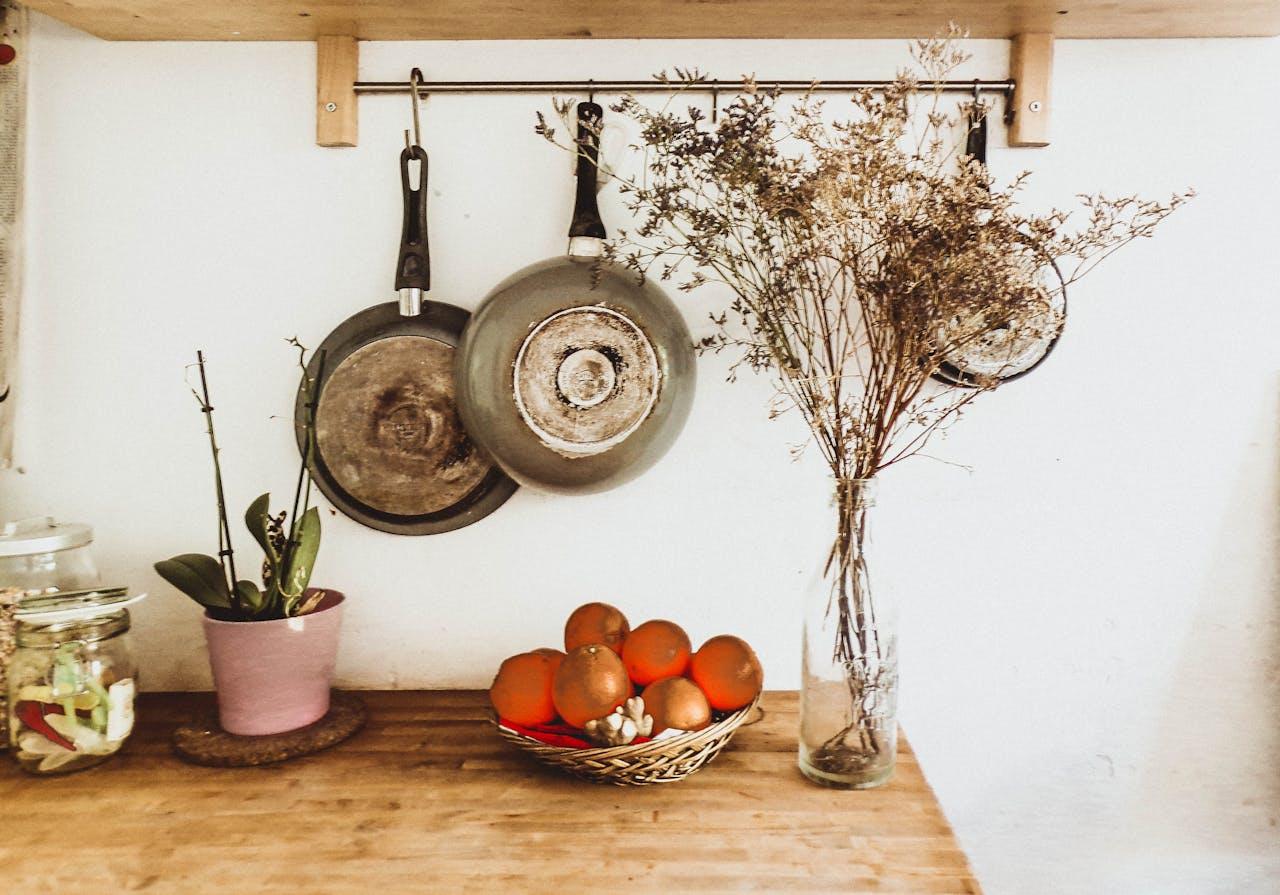 Rustic kitchen countertop with hanging pans, dried flowers, oranges in basket, and potted plant for home decor